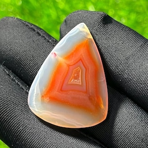 May include: A triangular polished agate gemstone featuring concentric bands of translucent white and vibrant orange-red colours. The stone has a smooth, glossy surface with a distinct central geometric pattern and a clear, crystalline core.