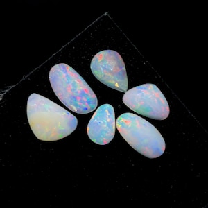 Puede incluir: Seis ópalos con base blanca lechosa y destellos iridiscentes de azul, rosa y verde. Las piedras preciosas, de formas ovaladas, de lágrima e irregulares, se exhiben sobre un fondo negro. Un conjunto de piedras preciosas naturales.