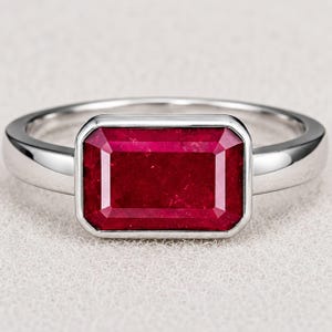 Könnte beinhalten: Ein silberfarbener Ring mit einem großen, rechteckigen, tiefroten Edelstein im Smaragdschliff, der in einer glatten Metallfassung sitzt. Die Ringschiene ist poliert und massiv, mit einem schlichten Design, das die leuchtende Farbe des Steins betont.