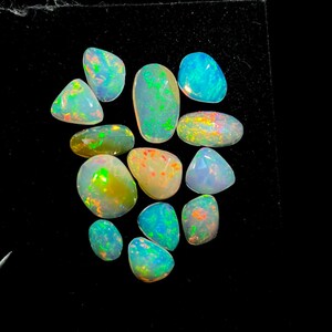 Puede incluir: Una colección de ópalos de diversas formas y tamaños, que muestra un vibrante juego de colores. Los ópalos exhiben una mezcla de azules, verdes, amarillos y naranjas, con algunas piedras translúcidas. Las piedras preciosas están sobre un fondo negro.