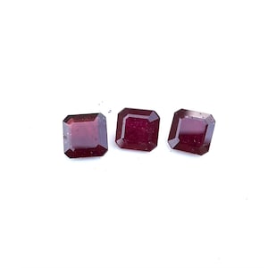 May include: Three rectangular, faceted gemstones in a deep red colour. The gems are arranged in a row against a white background, highlighting their polished surfaces and sharp edges. These gems are likely for jewellery making.