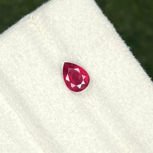 May include: A pear-shaped, faceted red gemstone with a brilliant cut. The stone displays a deep, saturated crimson hue and is positioned on a textured, off-white surface. The gemstone features multiple triangular facets that reflect light.