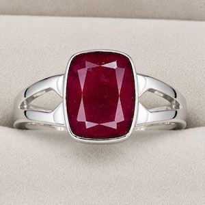 Puede incluir: Un anillo de tono plateado con una gran piedra preciosa de color rojo intenso en talla cojín, montada en una banda de metal pulido. La banda presenta un diseño de brazo dividido que se estrecha hacia el centro, enmarcando la piedra.