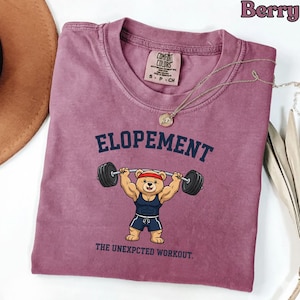Peut inclure: T-shirt de couleur baie avec le mot "ELOPEMENT" au-dessus d'un ours de dessin animé soulevant des poids. Le texte "THE UNEXPCCTED WORKOUT" est en dessous de l'ours. Une étiquette Comfort Colors est visible.