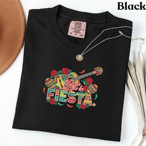 Puede incluir: Camiseta negra con un gráfico colorido de "Let's Fiesta" que presenta una guitarra, rosas y chiles. La camiseta tiene cuello redondo y mangas cortas. También se ve un collar plateado con un colgante.