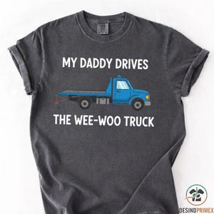 Könnte beinhalten: Dunkelgraues T-Shirt mit dem Text "MY DADDY DRIVES THE WEE-WOO TRUCK" über einer blauen Abschleppwagen-Grafik. Das Shirt hat hochgekrempelte Ärmel und einen Rundhalsausschnitt.