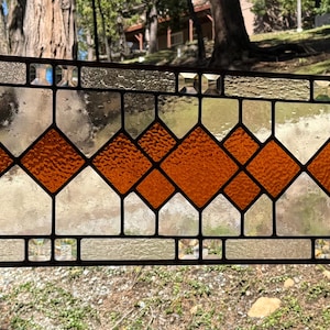 May include: Rectangular stained glass panel with a geometric design. The panel features clear, textured, and amber-colored glass pieces. The design includes diamond shapes and rectangular sections, all framed by a dark metal border. The panel is suspended by chains.