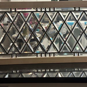 May include: Rectangular decorative mirror with a light wood frame. The mirror features a geometric design of diamond-shaped glass panes separated by black metal lines. The design is reminiscent of stained glass.