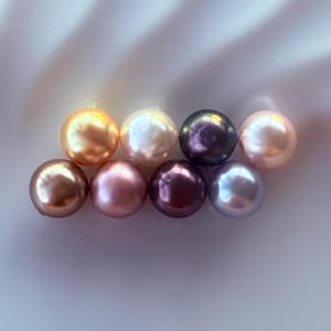 May include: A collection of eight round pearls in various colours, including white, cream, pink, purple, and brown. The pearls are arranged in a row on a white surface.
