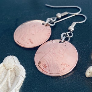 May include: A pair of copper penny earrings with silver hooks. The pennies are dated 2021 and feature the profile of Abraham Lincoln on one side and the Lincoln Memorial on the other.