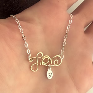 May include: A gold-toned necklace with a wire-wrapped "Love" pendant and a silver charm with the letter "R" hanging from it. The necklace is on a silver chain.