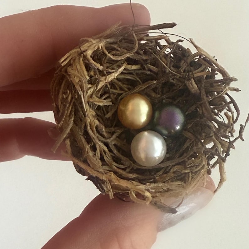May include: Three pearls nestled in a bird's nest. One pearl is gold, one is white, and one is iridescent purple.