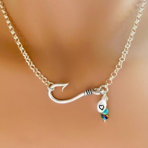 May include: A silver chain necklace with a silver fishing hook pendant and a small silver charm with a heart engraved on it. The charm is attached to three small iridescent beads.