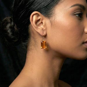 May include: A woman wearing a gold earring with a rectangular, amber-colored gemstone. The earring hangs from a gold hook. The woman's skin is a warm tone, and her hair is pulled back. The background is a dark, solid color.
