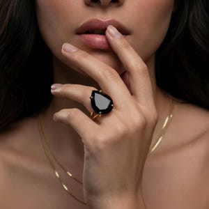 May include: A gold ring featuring a large, pear-shaped black gemstone. The ring is set on a delicate gold band. The image shows the ring on a person's finger, with a gold necklace in the background. The jewelry is elegant and eye-catching.