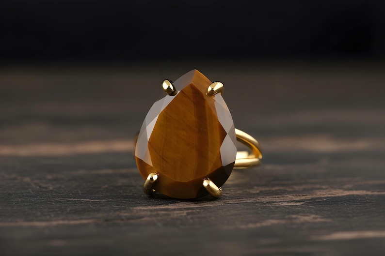 A large, oval-shaped amber-colored gemstone, possibly a diamond or amber, is displayed on a gold ring against a dark background.