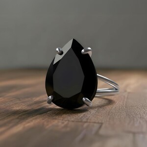 May include: A silver ring featuring a large, pear-shaped black gemstone. The ring's band is a split shank design, and the gemstone is held in place by four prongs. The ring is set against a wooden surface.