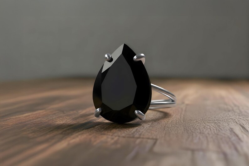 May include: A silver ring featuring a large, pear-shaped black gemstone. The ring's band is a split shank design, and the gemstone is held in place by four prongs. The ring is set against a wooden surface.