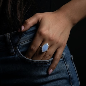 May include: A silver ring with a large, light blue, marquise-cut gemstone. The ring is set on a hand resting in a denim jeans pocket, against a dark background. The gemstone is the focal point.