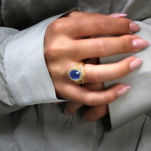 K&ouml;nnte beinhalten: Ein goldener Ring mit einem blauen ovalen Edelstein, der in einem dekorativen goldenen Rahmen gefasst ist. Der Ring wird am Zeigefinger einer Hand getragen.