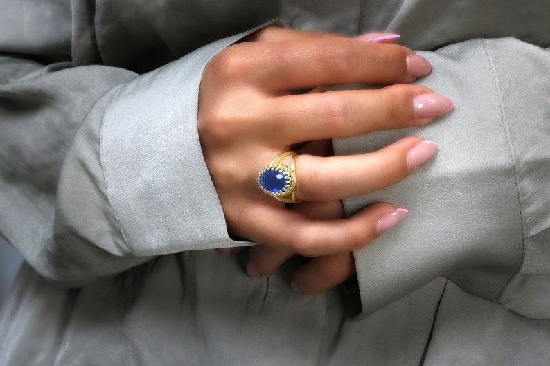 K&ouml;nnte beinhalten: Ein goldener Ring mit einem blauen ovalen Edelstein, der in einem dekorativen goldenen Rahmen gefasst ist. Der Ring wird am Zeigefinger einer Hand getragen.