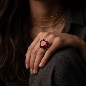 Op de afbeelding: Een opvallende ring met een grote, hartvormige, rode edelsteen. De ring is gezet op een zilveren band en wordt aan een vinger gedragen. Fijne gouden kettingen zijn zichtbaar. De afbeelding straalt luxe en stijl uit.