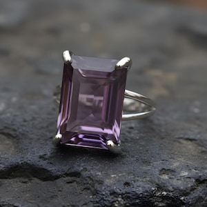 May include: A silver ring featuring a large, rectangular, faceted amethyst gemstone. The amethyst has a deep purple hue. The ring is set on a dark, textured surface, highlighting the gemstone's color and clarity. The ring's band is split.