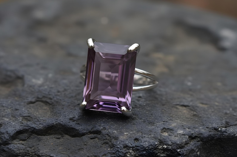 May include: A silver ring featuring a large, rectangular, faceted amethyst gemstone. The amethyst has a deep purple hue. The ring is set on a dark, textured surface, highlighting the gemstone's color and clarity. The ring's band is split.
