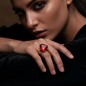 May include: A gold ring featuring a large, triangular, red gemstone. The ring is set on a hand, with the focus on the jewelry. The background is a blurred, dark color.