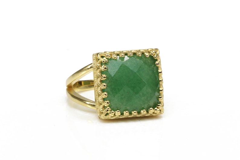 Puede incluir: Anillo dorado con una piedra preciosa verde cuadrada y facetada. El anillo tiene un dise&ntilde;o de banda dividida y un engaste decorativo de estilo filigrana alrededor de la piedra preciosa. Fondo blanco.