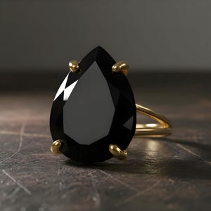 May include: A gold ring featuring a large, pear-shaped black gemstone. The gemstone is held in place by four gold prongs. The ring's band is a simple, polished gold. The ring is set against a dark, textured background.