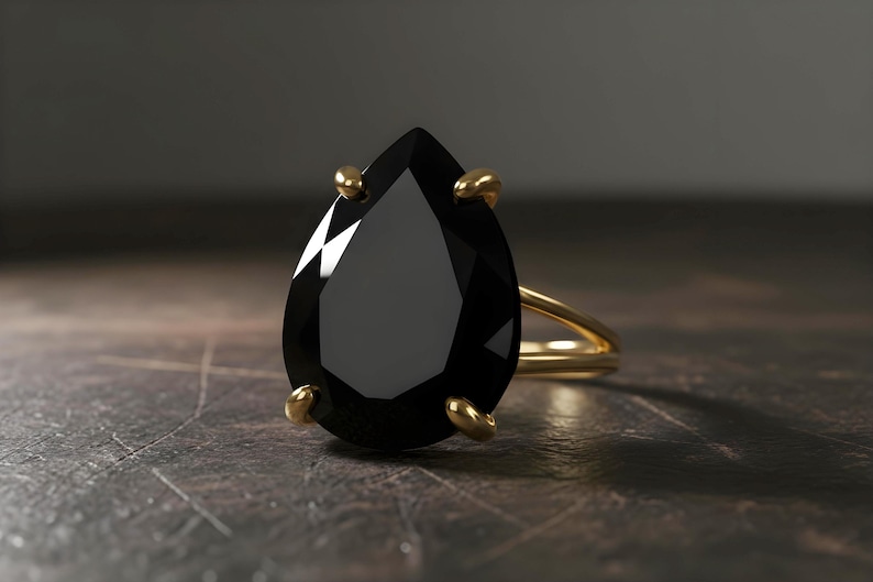 May include: A gold ring featuring a large, pear-shaped black gemstone. The gemstone is held in place by four gold prongs. The ring's band is a simple, polished gold. The ring is set against a dark, textured background.