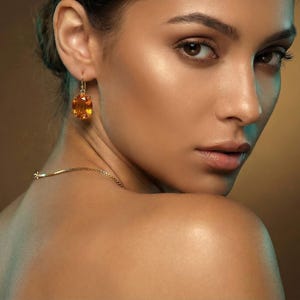 May include: A woman wearing a gold necklace and earrings with large, oval, amber-colored gemstones. The earrings are suspended from gold hooks. The woman's skin has a warm, golden tone, and she is looking over her shoulder.