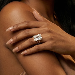 May include: A large, emerald-cut diamond ring with a split shank setting. The ring's clear gemstone is set in a silver band. The ring is displayed on a hand with long, polished nails. The skin tone is dark.