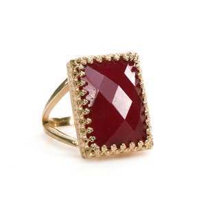 May include: A gold-toned ring featuring a large, rectangular, faceted, deep red gemstone. The gemstone is set in a decorative gold frame with a scalloped edge. The ring has a split shank design.