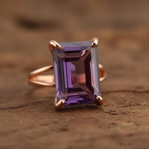 May include: A rose gold ring featuring a large, rectangular, faceted amethyst gemstone. The amethyst has a deep purple hue with lighter shades within. The ring's band has a split design, and the gemstone is held in place by four prongs.