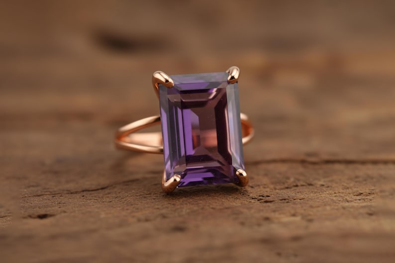 May include: A rose gold ring featuring a large, rectangular, faceted amethyst gemstone. The amethyst has a deep purple hue with lighter shades within. The ring's band has a split design, and the gemstone is held in place by four prongs.