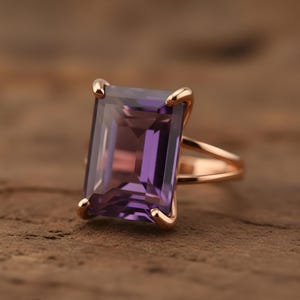 May include: Rose gold ring featuring a large, rectangular, purple amethyst gemstone. The gemstone is set with four prongs. The ring's band has a split shank design. The ring is displayed on a textured, brown surface.