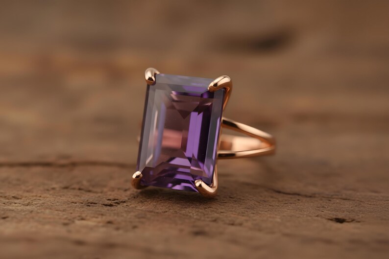 May include: Rose gold ring featuring a large, rectangular, purple amethyst gemstone. The gemstone is set with four prongs. The ring's band has a split shank design. The ring is displayed on a textured, brown surface.