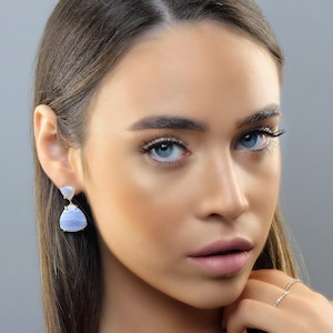 Puede incluir: Primer plano de una persona que lleva pendientes de color azul claro y dorado. Los pendientes tienen forma de lágrima con una piedra más pequeña en la parte superior. La persona tiene ojos azules y lleva un anillo en el dedo.