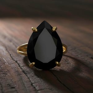 May include: A gold ring with a large, pear-shaped black gemstone. The ring's band is a simple, double-band design. The gemstone is held in place by four gold prongs. The ring is set against a dark wooden surface.