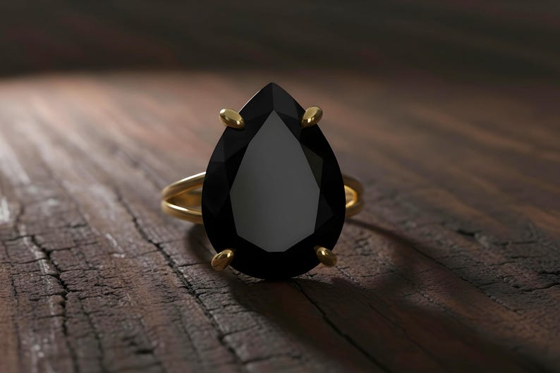 May include: A gold ring with a large, pear-shaped black gemstone. The ring's band is a simple, double-band design. The gemstone is held in place by four gold prongs. The ring is set against a dark wooden surface.