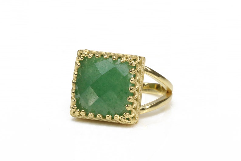 Puede incluir: Anillo dorado con una piedra preciosa verde cuadrada y facetada. La piedra preciosa est&aacute; engastada en un marco decorativo calado. El anillo tiene un dise&ntilde;o de ca&ntilde;a dividida.