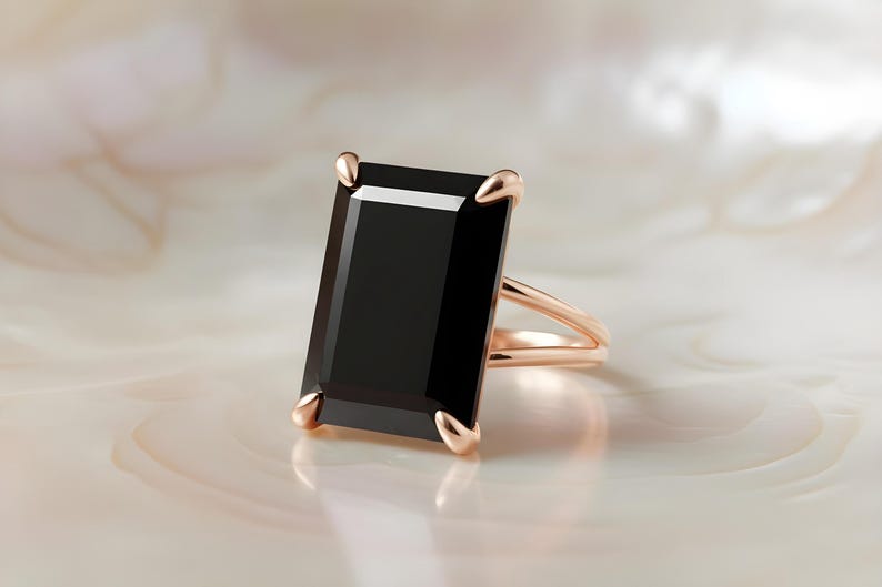 Puede incluir: Un anillo de oro rosa con una gran piedra negra rectangular. El anillo tiene un dise&ntilde;o de ca&ntilde;a dividida y cuatro garras que aseguran la piedra. El engaste y la banda tienen un acabado pulido, sobre un fondo neutro.