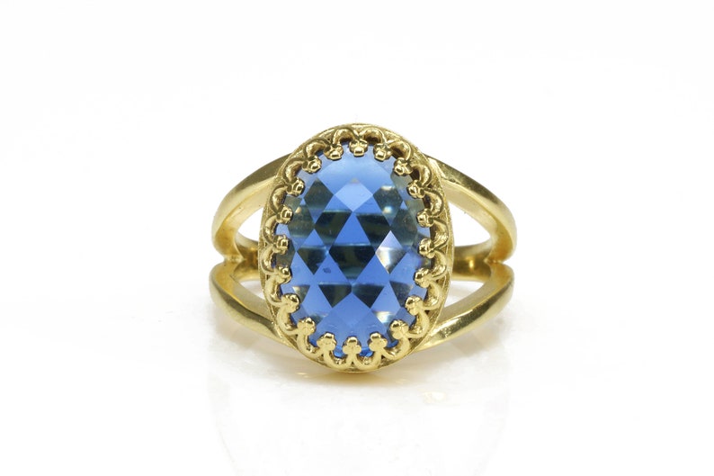 K&ouml;nnte beinhalten: Ein goldener Ring mit einem blauen, ovalen Edelstein, der in einem dekorativen Goldrahmen gefasst ist.