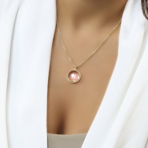 Op de afbeelding: Een gouden ketting met een roze edelsteen hanger. De hanger is rond en heeft een delicate, parelrand.