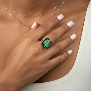 May include: A gold ring with a large, emerald-cut green gemstone. The ring is set in a gold band and is worn on a finger. A delicate gold necklace is also visible. The person is wearing a white vest top.