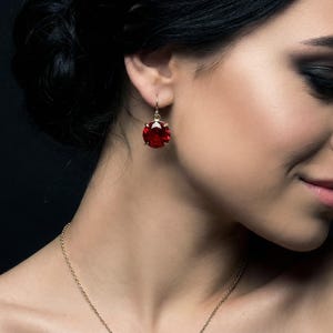 May include: A gold drop earring featuring a round, faceted deep red gemstone suspended from a thin gold hook. The earring is paired with a matching gold chain necklace that rests against the collarbone, showcasing a polished metal finish.