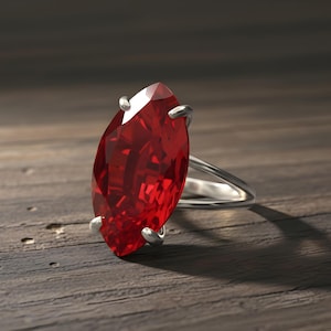 May include: A marquise-cut, ruby-red gemstone ring with a silver band. The gemstone is held in place by four silver prongs. The ring sits on a wooden surface.