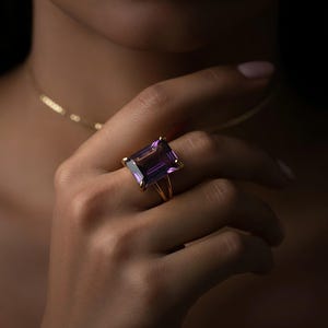 May include: A gold ring featuring a large, rectangular, purple amethyst gemstone. The ring is set on a gold band with a split shank design. The ring is worn on a hand, with a gold necklace visible in the background.
